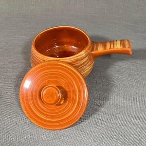 Könnte beinhalten: Eine braune Keramik-Auflaufform mit Deckel. Die Form hat ein Wirbelmuster in Orange und Gelb. Der Deckel hat einen Knopf oben.