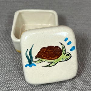 May include: A small, open, cream-coloured ceramic trinket box with a square lid. The lid features a hand-painted sea turtle with a brown shell and green body, surrounded by blue water and seaweed. The box has a yellow rim.