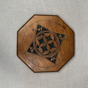 May include: Octagonal wooden coaster with a carved geometric design in the centre. The design is dark brown and features a star shape with a circle inside.