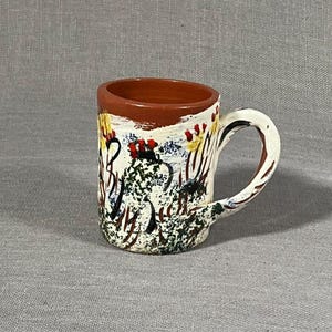 Może przedstawiać: Mały, biały kubek ceramiczny z brązową krawędzią i uchwytem. Kubek jest ozdobiony kolorowym abstrakcyjnym wzorem z czarnej, zielonej, żółtej, czerwonej i niebieskiej farby.
