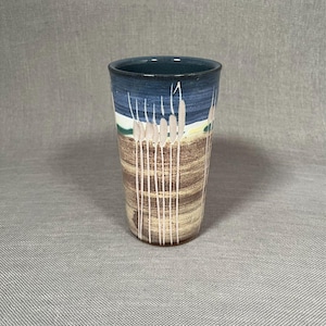 May include: A ceramic mug with a blue, brown, white, and green design. The mug has a tall, cylindrical shape and features a pattern of vertical lines and abstract shapes.