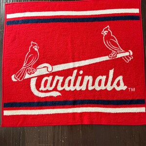 May include: Red and white throw blanket with the St. Louis Cardinals logo. The logo features two white cardinals perched on a white baseball bat with the word "Cardinals" in white cursive script and a trademark symbol.