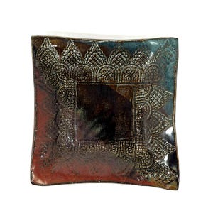 Può includere: Piatto quadrato in ceramica con un motivo decorativo a forma di pizzo. Il piatto presenta una sfumatura di colori, tra cui rosso, marrone e blu. Il design intricato è in rilievo, creando una superficie strutturata.