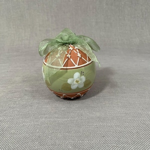 Peut inclure: Une décoration d'œuf de Pâques en céramique peinte à la main, de couleur verte et orange, avec des fleurs blanches et un ruban vert.