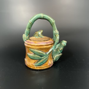 May include: A small ceramic teapot with a frog on the lid and a bamboo design. The teapot is glazed in shades of green and brown.
