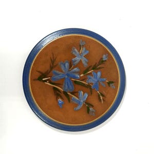 May include: A round ceramic coaster with a blue rim and a brown center. The coaster features a hand-painted design of blue flowers with green stems.