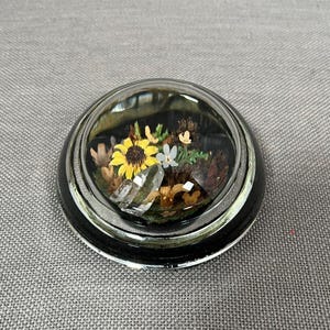 May include: A small, round, glass dome with a black rim sits on a gray fabric surface. Inside the dome, there are dried flowers and a clear crystal.