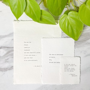 以下が含まれることがあります： 大理石の表面に配置された、印刷された引用句が書かれた3枚の手作りの紙。背景には緑の葉があります。引用句は黒インクで書かれており、「You are the finest」や「She was an adventurer」などのフレーズが含まれています。