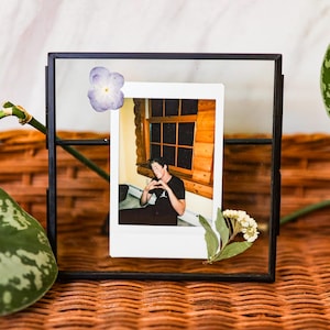May include: A black metal and glass photo frame holding a Polaroid-style photo. The photo features a person making a heart shape with their hands. Dried flowers and a purple flower are attached to the frame.