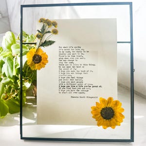 May include: A framed print with a quote by Francis Scott Fitzgerald, decorated with dried yellow sunflowers and other small yellow flowers. The frame is black and the background is a light beige color.