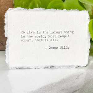 May include: A white rectangular card with a quote by Oscar Wilde, "To live is the rarest thing in the world. Most people exist, that is all." The card has a textured, torn edge. Green leaves and a brown pot are in the background.