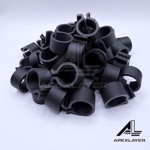 Può includere: Un mucchio di clip di plastica nera, ciascuna con un corpo circolare e un'apertura curva. Le clip sono di varie dimensioni e forme e sono sparse su uno sfondo bianco. Il logo "APEXLAYER" si trova nell'angolo in basso a destra.