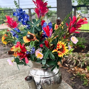 May include: A vibrant floral arrangement in a silver teapot. The bouquet features red, blue, yellow, and pink flowers with green foliage. The teapot has a spout, handle, and decorative embossed details. The arrangement is set on a concrete surface.