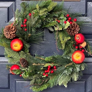 May include: A circular wreath constructed from green pine needles and holly leaves, decorated with brown pinecones, bright red artificial apples, and clusters of small red berries. The arrangement is dense and features varied textures of foliage.