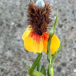 May include: A vibrant artificial flower with a yellow and orange petal design. The flower has a spiky, brown and purple center with a light blue top. Green leaves and stem complete the floral arrangement.