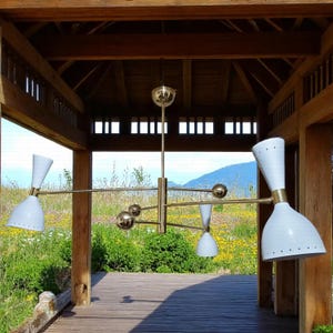 May include: A mid-century modern chandelier with a brass finish and white cone-shaped shades. The fixture has multiple arms extending from a central stem, with spherical accents. The chandelier is suspended from a wooden structure, with a natural outdoor background.