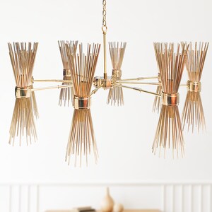 May include: A gold-toned, mid-century modern chandelier with multiple light fixtures. The chandelier features a central stem with radiating arms, each holding a cluster of thin, vertical rods. The design is symmetrical and creates a starburst effect, suitable for interior lighting.