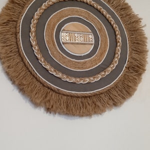 Peut inclure: Décoration murale circulaire d'esthétique bohème naturelle. Elle présente un cercle central en bois avec un motif géométrique, entouré d'anneaux concentriques en tissu gris, corde et jute frangée. La palette de couleurs globale est neutre.