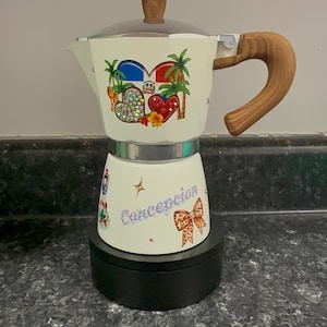 May include: Cream-colored stovetop espresso maker with a wooden handle and knob. The design includes colorful heart and palm tree graphics, along with the word "Concepcion" and a bow.