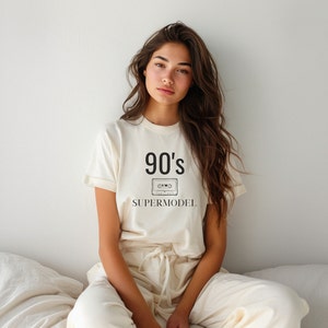 Può includere: Una donna indossa una maglietta bianca con il testo "90's SUPERMODEL" e una grafica di una cassetta. È seduta su un letto bianco con un cuscino bianco.