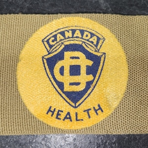 Puede incluir: Un parche de tela beige con un emblema circular amarillo que presenta un diseño de escudo azul y las palabras "CANADA HEALTH". El parche tiene forma rectangular con esquinas anguladas.