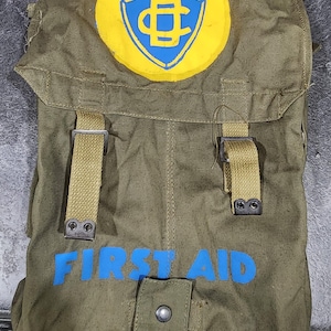 Puede incluir: Botiquín de primeros auxilios de lona verde oliva con las palabras "FIRST AID" en letras azules. Un emblema amarillo y azul con la palabra "CANADA" está en la parte superior. El kit tiene dos correas y un cierre de solapa.