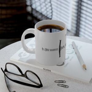 May include: White ceramic mug filled with dark coffee, featuring the text "As iron sharpens iron". The mug sits on a book with a pencil and eyeglasses nearby. The mug has a curved handle.