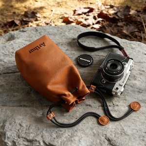 May include: A brown leather camera bag with the name "Arthur" embossed on the front. A black and silver camera with a lens cap and strap are next to the bag. The camera is on a gray rock.