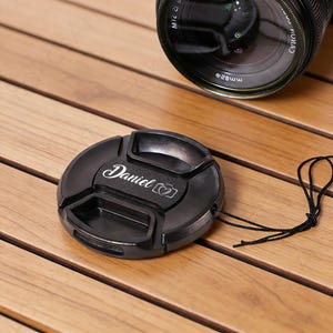 May include: A black camera lens cap with the name "Daniel" in cursive script and a camera icon. A black lens is partially visible in the background. The cap has a black string attached.