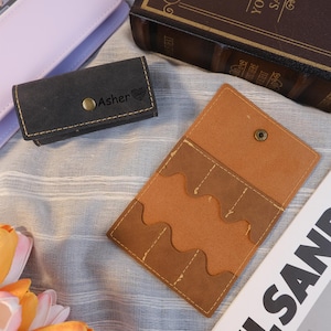 May include: A black leather cardholder with the name "Asher" and a heart symbol embossed on the front. The cardholder is closed with a snap. An open brown leather wallet with multiple card slots is also visible.