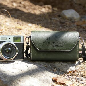 May include: A vintage camera next to a dark green leather camera bag with the text "Anna's CAMERA" in a script font. The bag has a white stitched border and a shoulder strap. The camera is black and silver.