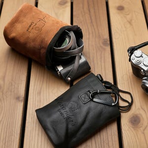 May include: Two leather camera cases, one brown and one black, sit on a wooden surface. The brown case, labeled "Anna's Camera," is open, revealing a camera and lens. The black case, labeled "Alexander's Camera," has a drawstring and carabiner.