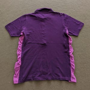 May include: A purple short-sleeved polo shirt with a contrasting light purple ruffled panel on the sides. The shirt has a classic collar and is made of a soft fabric. The back of the shirt is visible.
