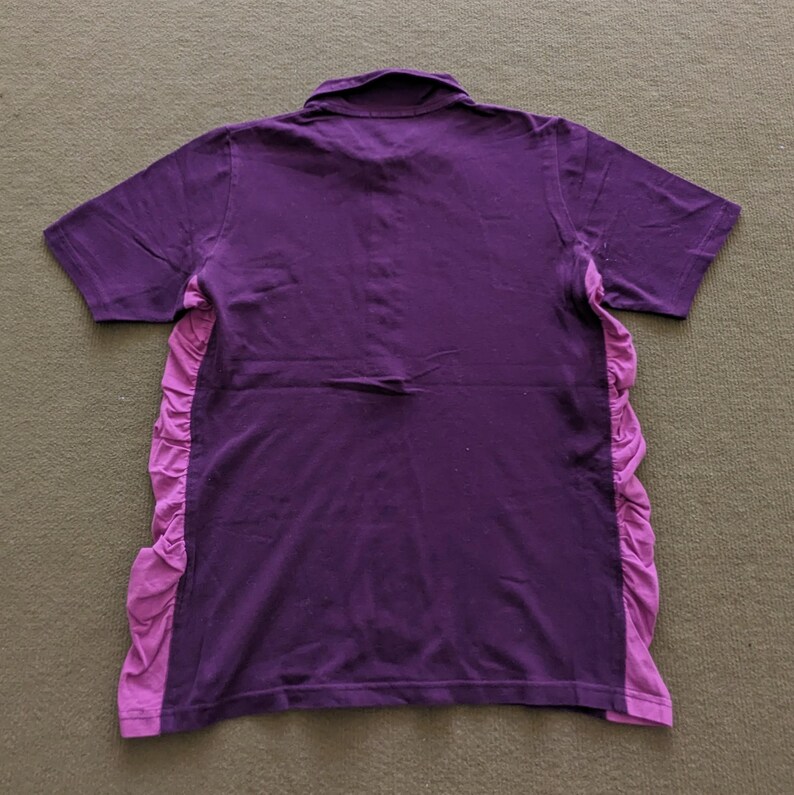 May include: A purple short-sleeved polo shirt with a contrasting light purple ruffled panel on the sides. The shirt has a classic collar and is made of a soft fabric. The back of the shirt is visible.