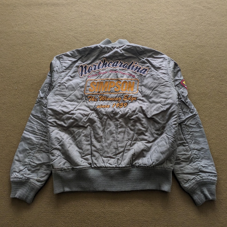 May include: A grey bomber jacket with "North Carolina" and "Simpson" embroidered on the back. The jacket has a ribbed collar, cuffs, and waistband.