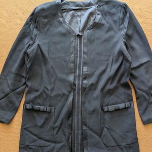 May include: A long black blazer with a V-neck and two pockets. The blazer has a satin trim down the front and a bow detail on each pocket.