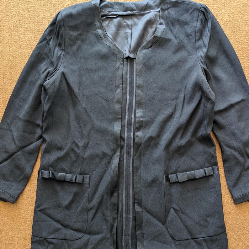 May include: A long black blazer with a V-neck and two pockets. The blazer has a satin trim down the front and a bow detail on each pocket.