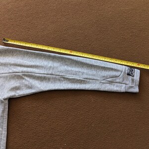 以下が含まれることがあります： グレーのスウェットシャツの袖に、黒いタグが付いていて、「ODD」と書かれています。袖は黄色のメジャーで測定されています。