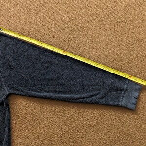 Op de afbeelding: Een donkergrijs longsleeve shirt met een geribbelde textuur. De mouw wordt gemeten met een gele meetlint, wat een lengte van 66 cm laat zien.