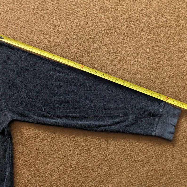 Op de afbeelding: Een donkergrijs longsleeve shirt met een geribbelde textuur. De mouw wordt gemeten met een gele meetlint, wat een lengte van 66 cm laat zien.