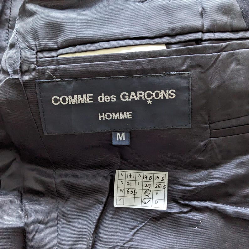 Puede incluir: Primer plano del interior de una prenda negra, que muestra una etiqueta con "COMME des GAR&Ccedil;ONS HOMME" en texto blanco. Una etiqueta de talla "M" y una etiqueta m&aacute;s peque&ntilde;a con medidas tambi&eacute;n son visibles. La tela tiene un ligero brillo.