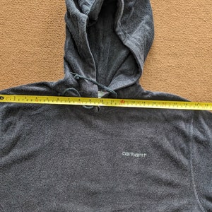 Op de afbeelding: Een donkergrijze hoodie met een Carhartt-logo op de voorkant. De hoodie ligt plat op een bruine ondergrond met een meetlint over de borst. Het lint meet 50,8 cm.