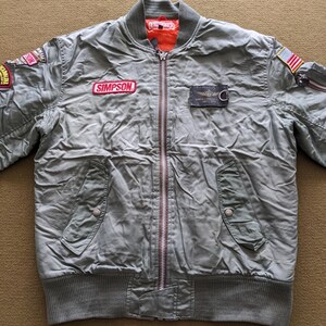 May include: A green bomber jacket with a zip fastening and multiple patches, including a "SIMPSON" patch, a "WHITE LINE" patch, and a US flag patch.