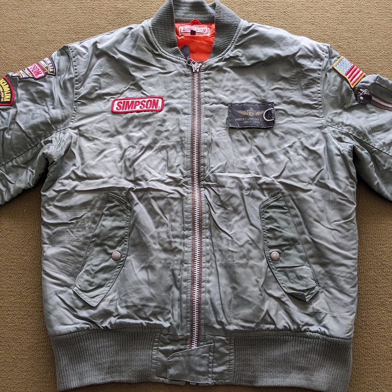 May include: A green bomber jacket with a zip fastening and multiple patches, including a "SIMPSON" patch, a "WHITE LINE" patch, and a US flag patch.