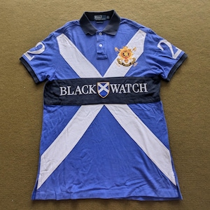 May include: A blue polo shirt with a white and blue Scottish tartan pattern. The shirt has a black band across the chest with the text "BLACK WATCH" and a Scottish flag emblem. The shirt has a small embroidered logo on the left chest.