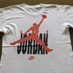 May include: White t-shirt with a red and black graphic of a basketball player jumping with the word "JORDAN" in black letters and "NIKE" in red letters below.