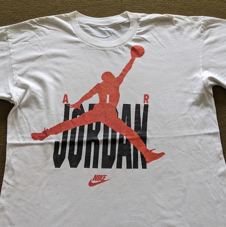 May include: White t-shirt with a red and black graphic of a basketball player jumping with the word "JORDAN" in black letters and "NIKE" in red letters below.