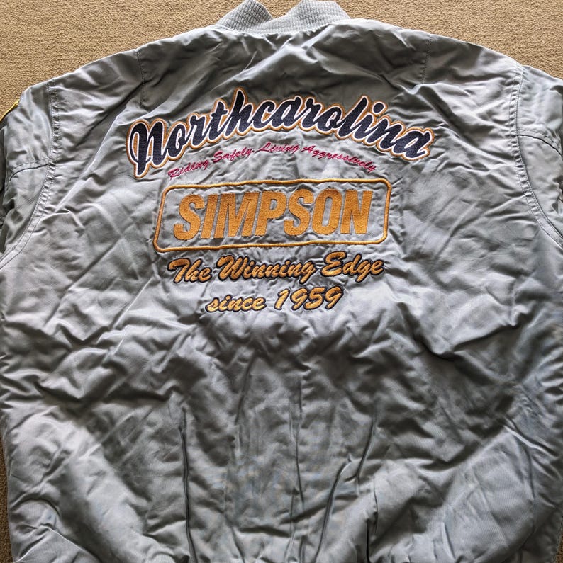 May include: A light blue bomber jacket with a yellow embroidered logo that says "North Carolina" and "Simpson The Winning Edge since 1959".