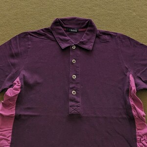 May include: A dark purple polo shirt with a button-down placket and a collared neckline. The shirt has short sleeves and a contrasting pink fabric panel on the side. The text "vf for living" is visible on the collar.