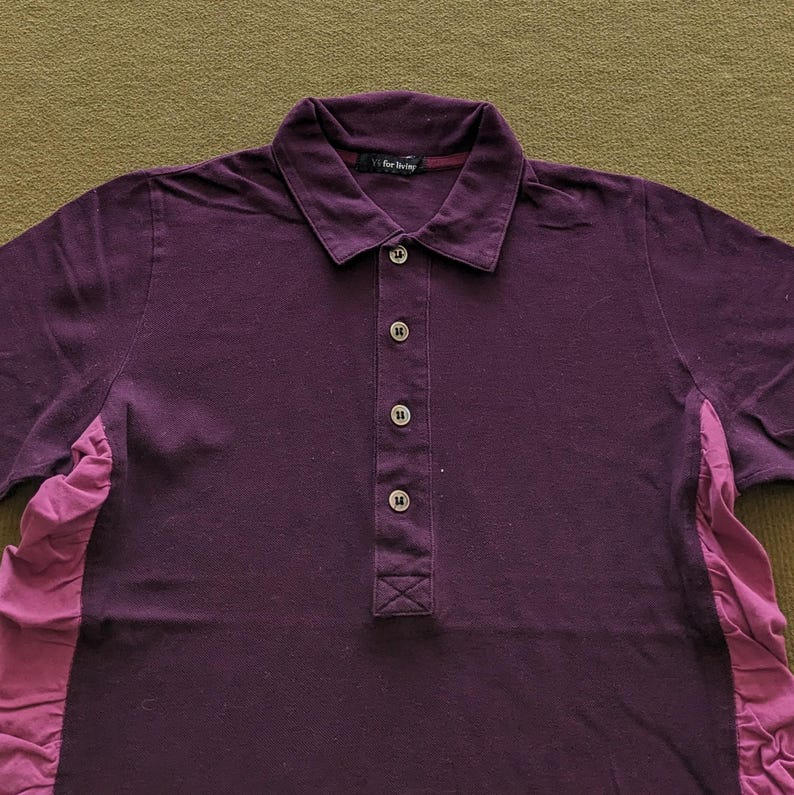 May include: A dark purple polo shirt with a button-down placket and a collared neckline. The shirt has short sleeves and a contrasting pink fabric panel on the side. The text "vf for living" is visible on the collar.